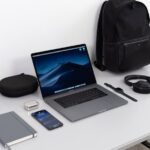 Essentials - MacBook Pro on table near black smartphone, cordless headphones and backpack