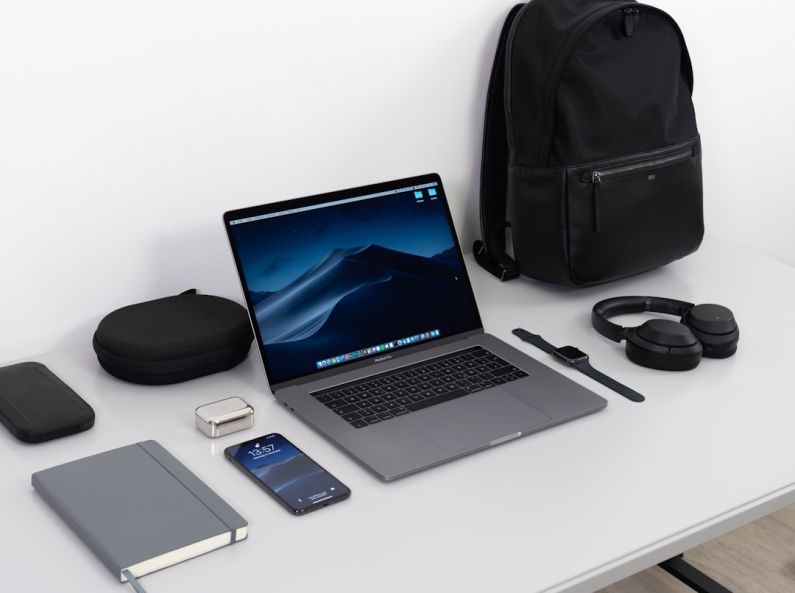 Essentials - MacBook Pro on table near black smartphone, cordless headphones and backpack
